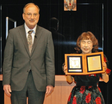 キサラ学長（左）から表彰盾を渡され笑顔になった青木さん＝名古屋市昭和区の南山大で