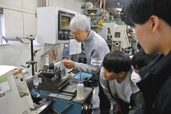 金属を削りバチの形に仕上がっていく様子を見守る学生たち＝亀山市能褒野町のギルドデザイン本社で